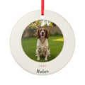 Pet photo ceramic Christmas ornament mock-up with a sample spaniel photo on a lawn, '2026' and 'Rufus', on a red ribbon
