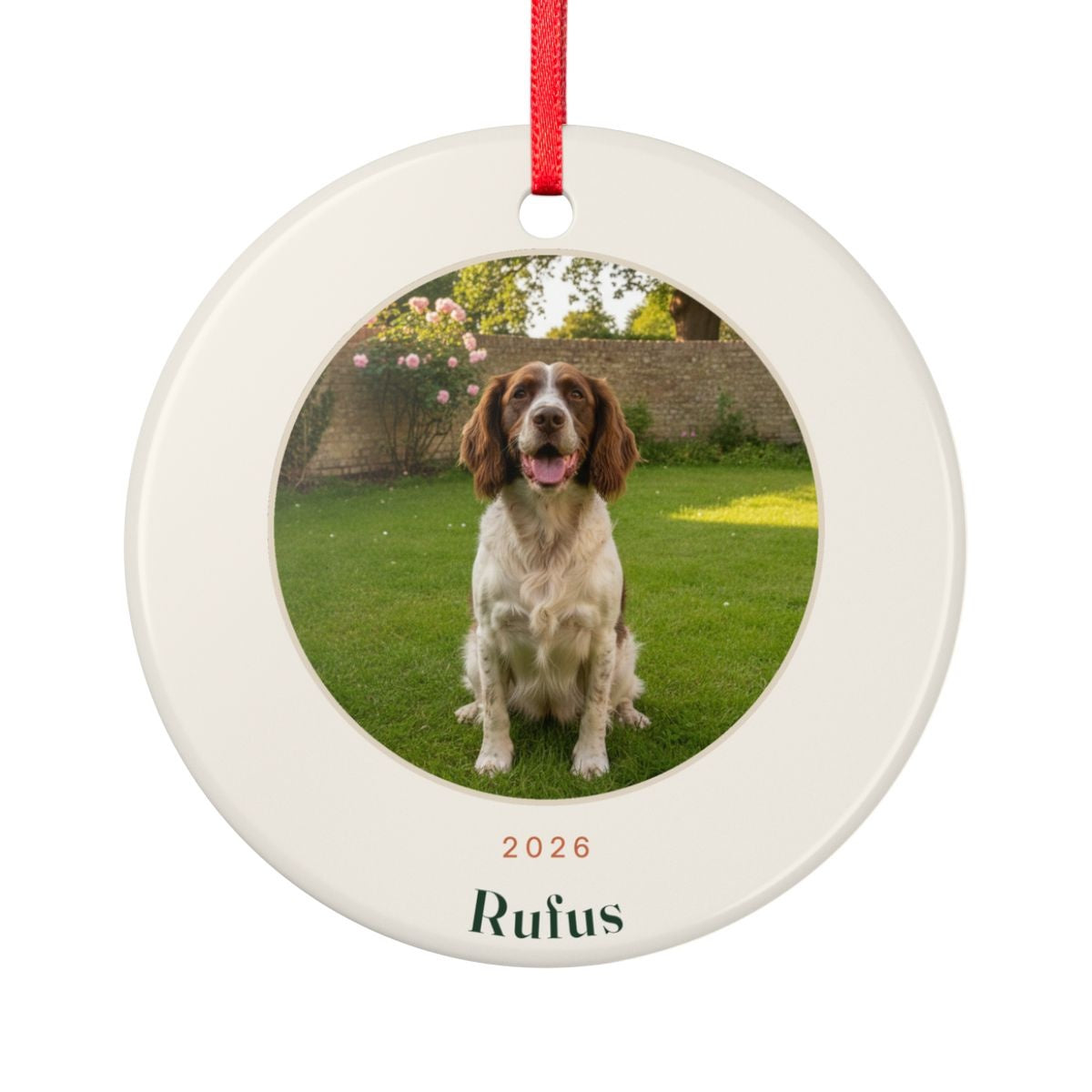 Pet photo ceramic Christmas ornament mock-up with a sample spaniel photo on a lawn, '2026' and 'Rufus', on a red ribbon