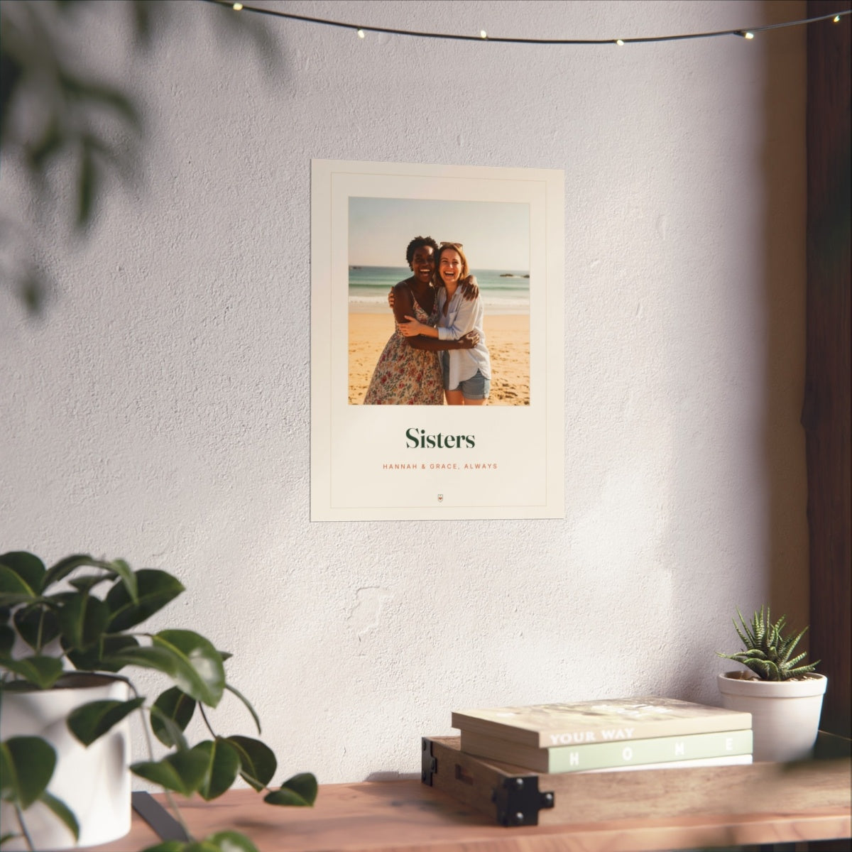Sisters photo print mock-up on a white wall above a desk with books, plants and fairy lights, larger size