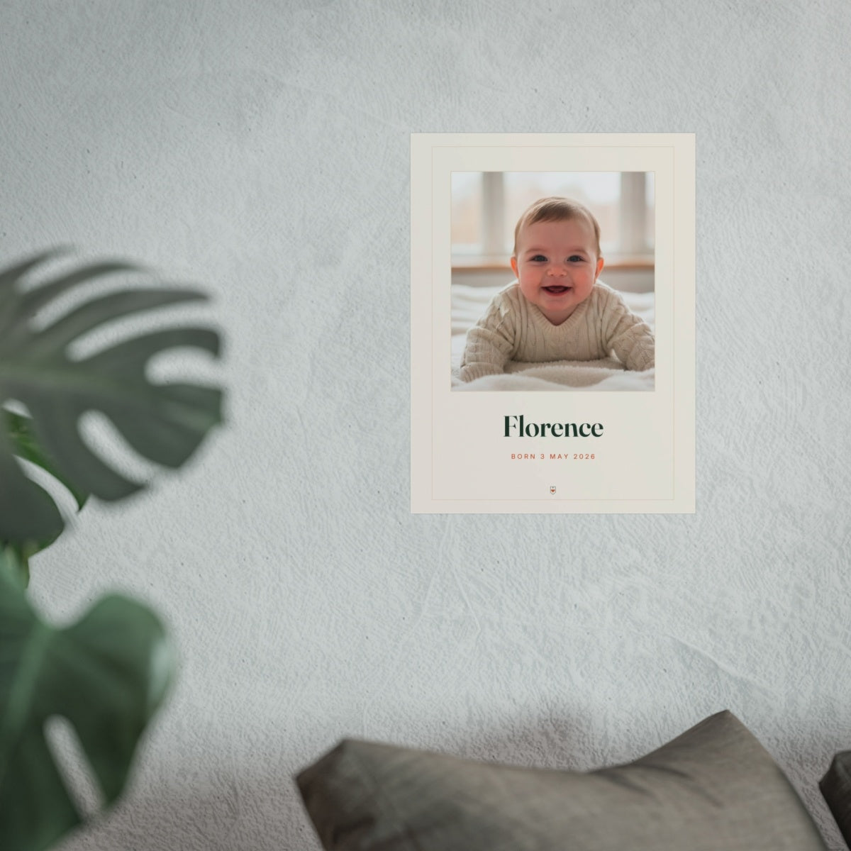 Baby photo print mock-up hung on a pale wall beside a monstera plant and a grey sofa, shown at the larger size