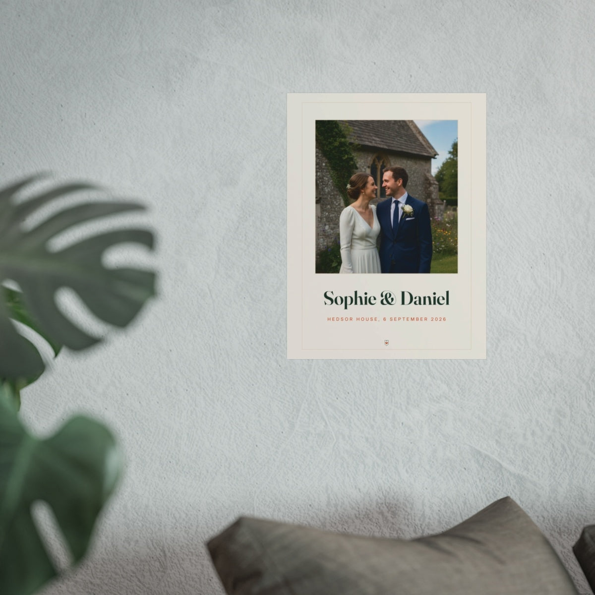 Wedding photo print mock-up hung on a pale textured wall above a grey sofa, with a monstera leaf in the foreground
