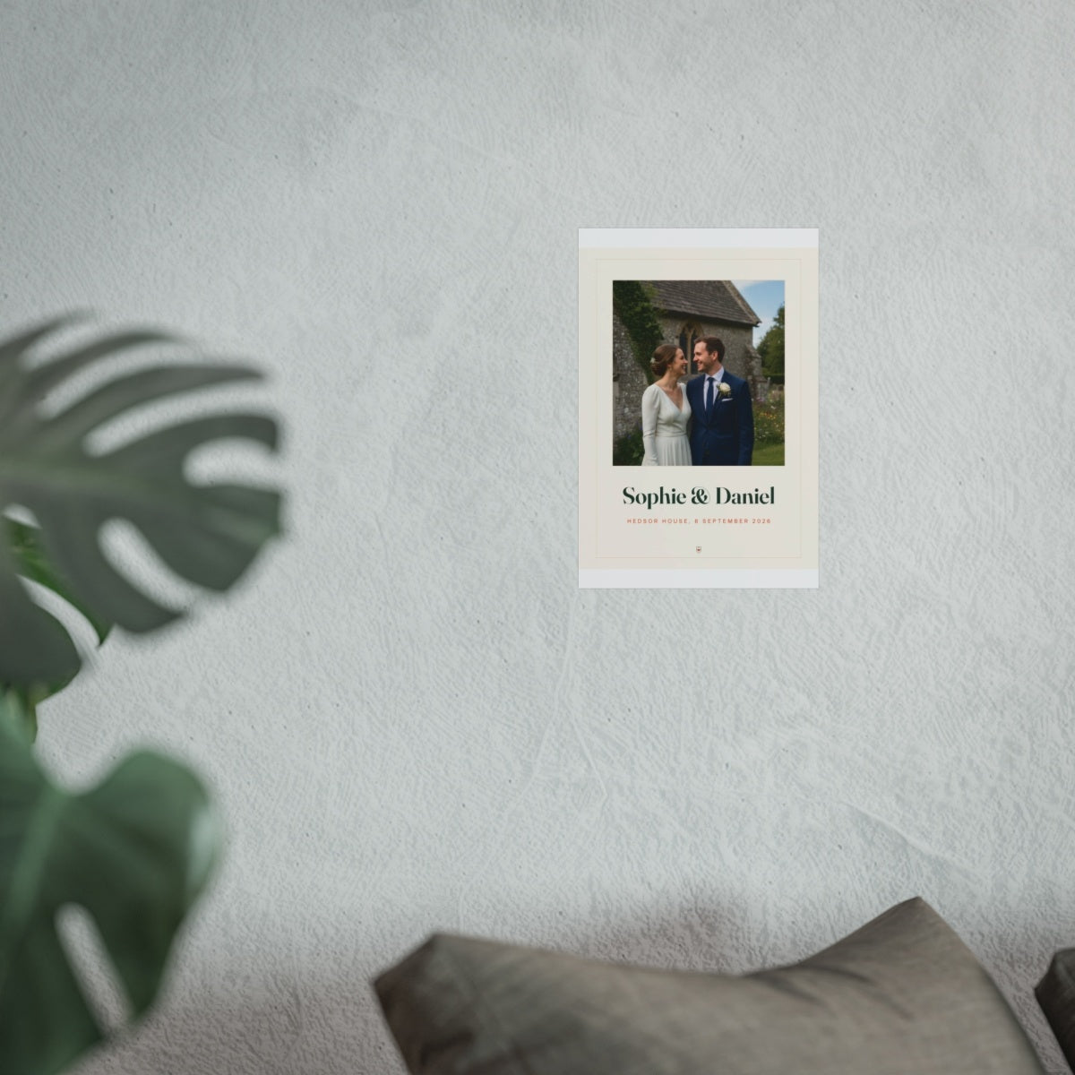 Wedding photo print mock-up shown smaller on the pale wall above the grey sofa, monstera leaf in front