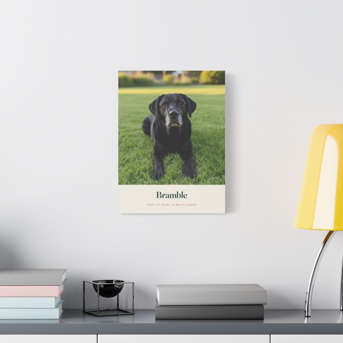 Pet memorial photo canvas mock-up on a white wall above a desk with books and a yellow lamp, Labrador photo in view