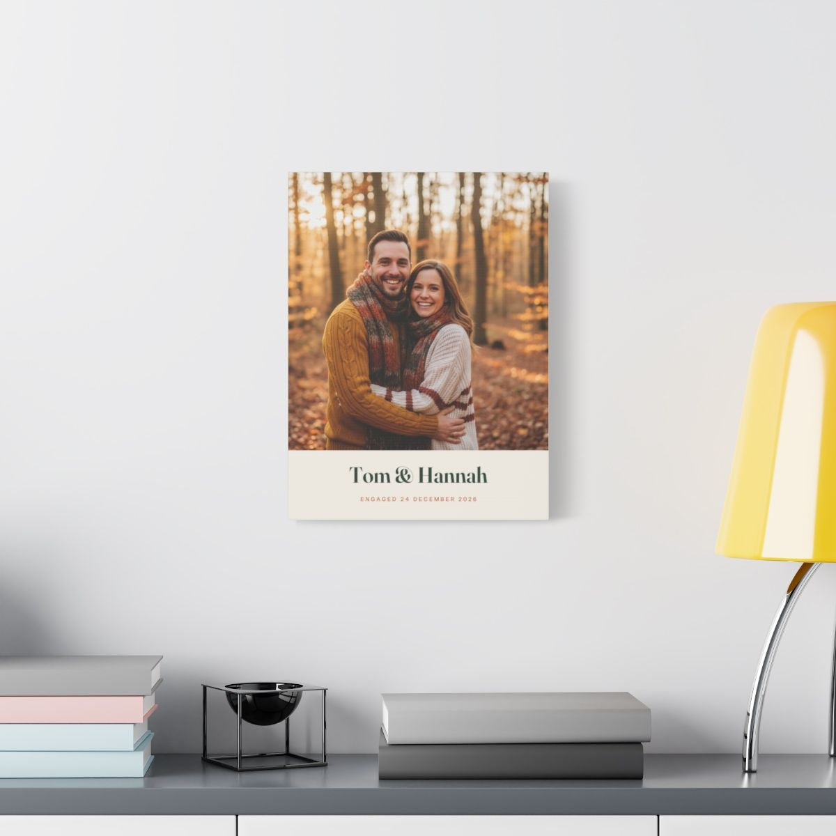 Engagement photo canvas hung on a white wall above a shelf with books and a yellow lamp, sample couple photo