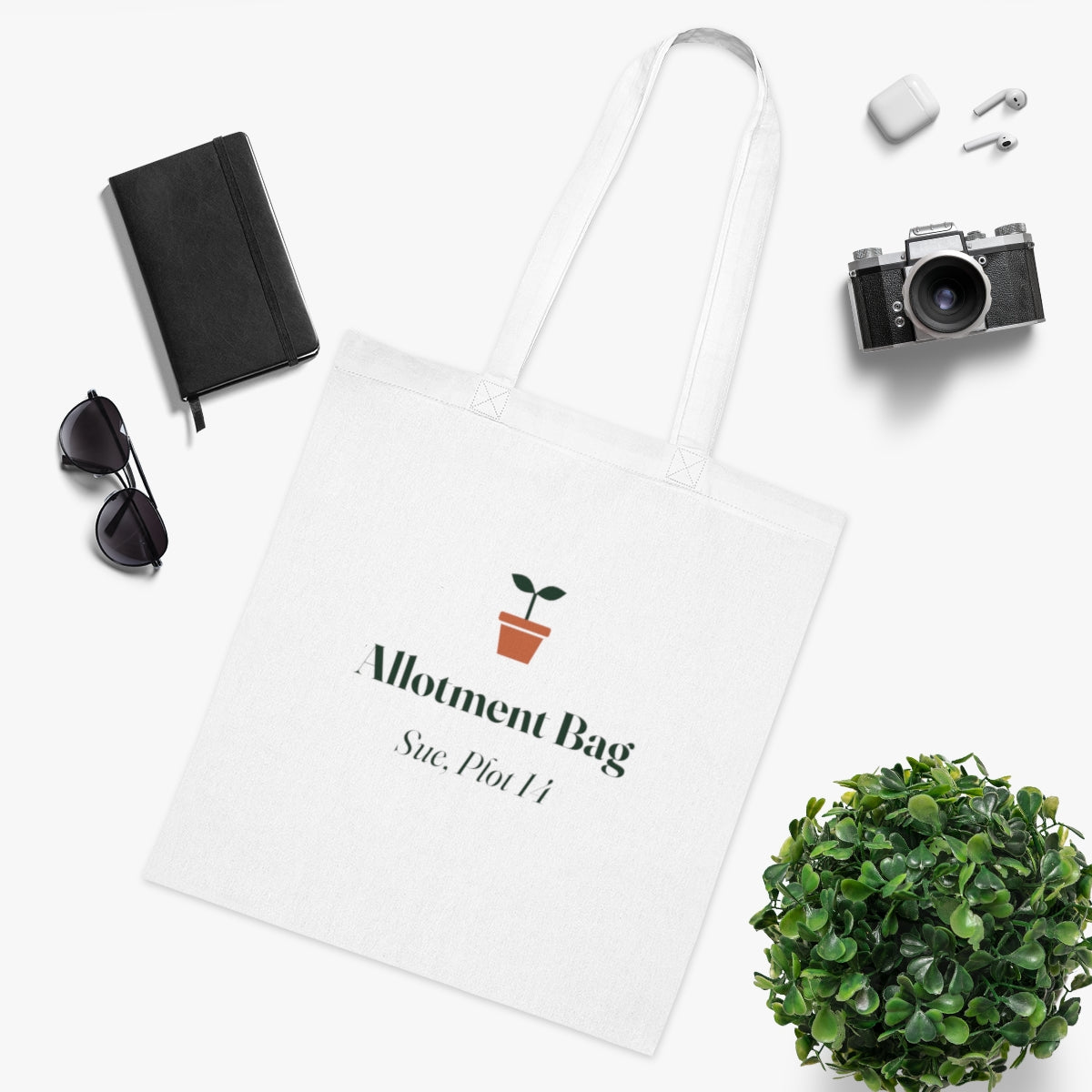 Allotment tote bag mock-up in a flat lay with a notebook, sunglasses, camera, earbuds and a leafy plant
