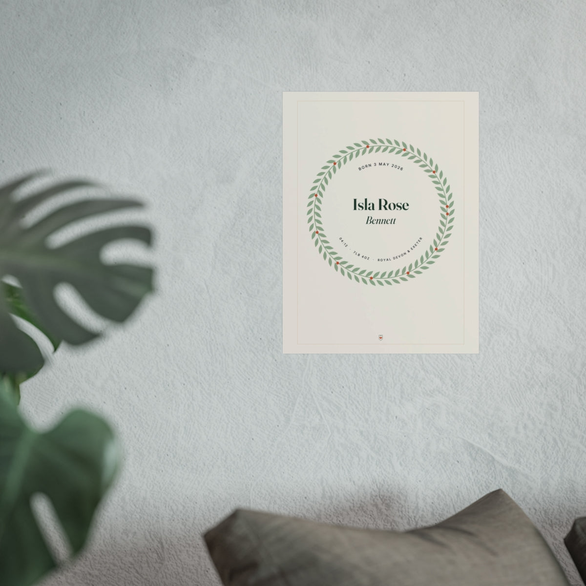 Birth details print mock-up on a white wall beside a monstera plant and a grey sofa cushion, 'Isla Rose Bennett' in a wreath