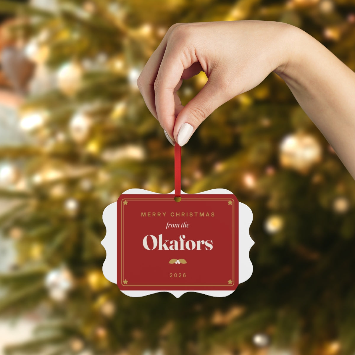 Red family aluminium plaque ornament with the example name the Okafors, held up on a ribbon against a Christmas tree