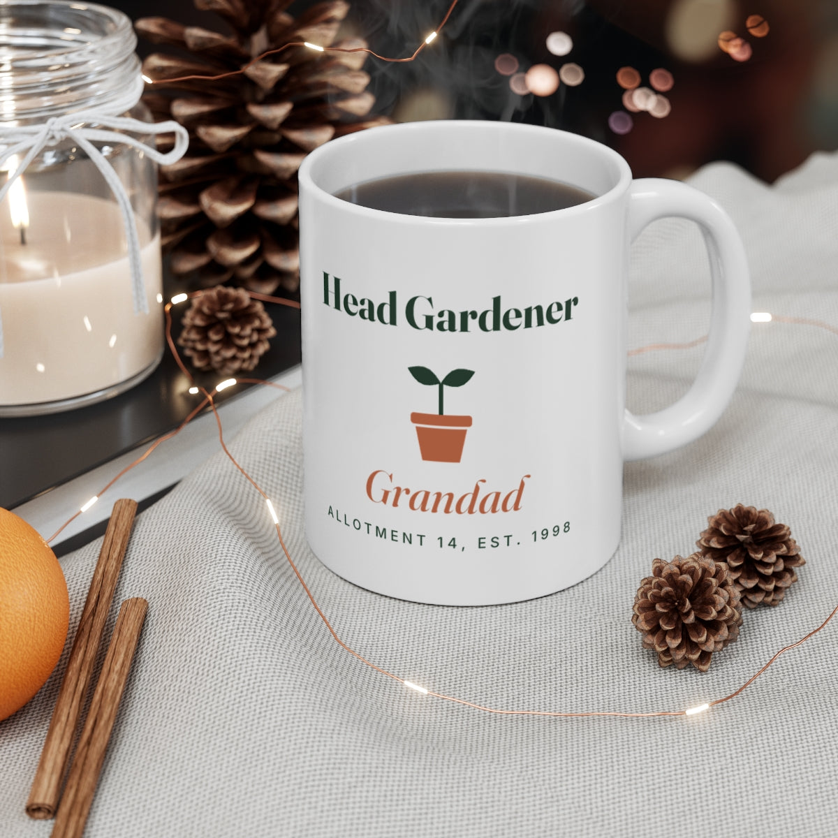 Head Gardener mug mock-up filled with coffee on a table with a candle, pine cones, cinnamon sticks and fairy lights