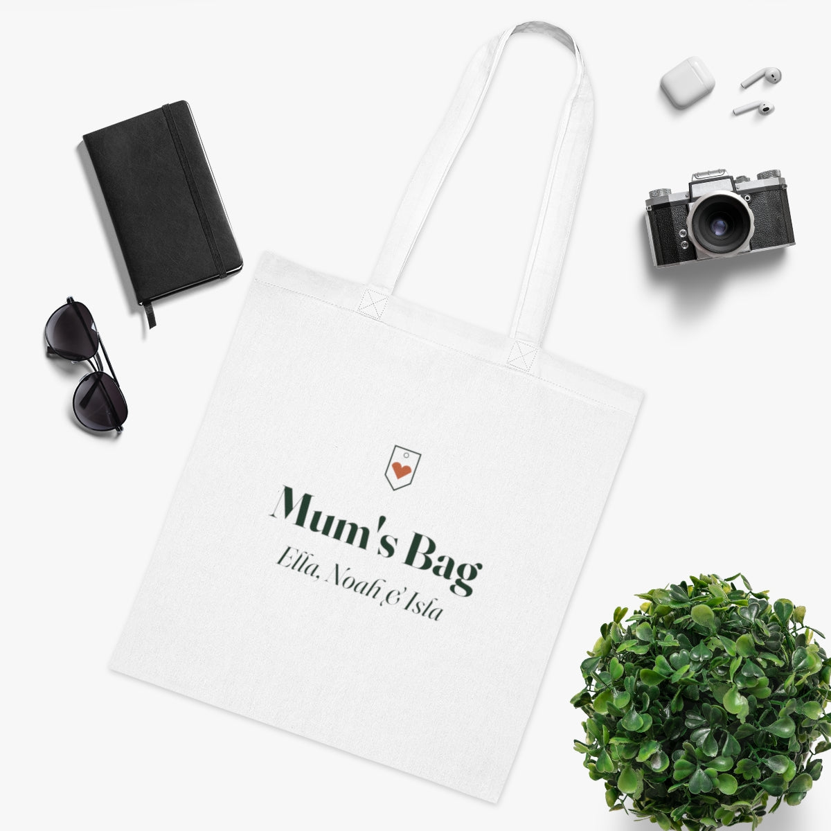 Personalised Mum's Bag tote mock-up laid flat beside a notebook, sunglasses, earbuds, a camera and a leafy plant