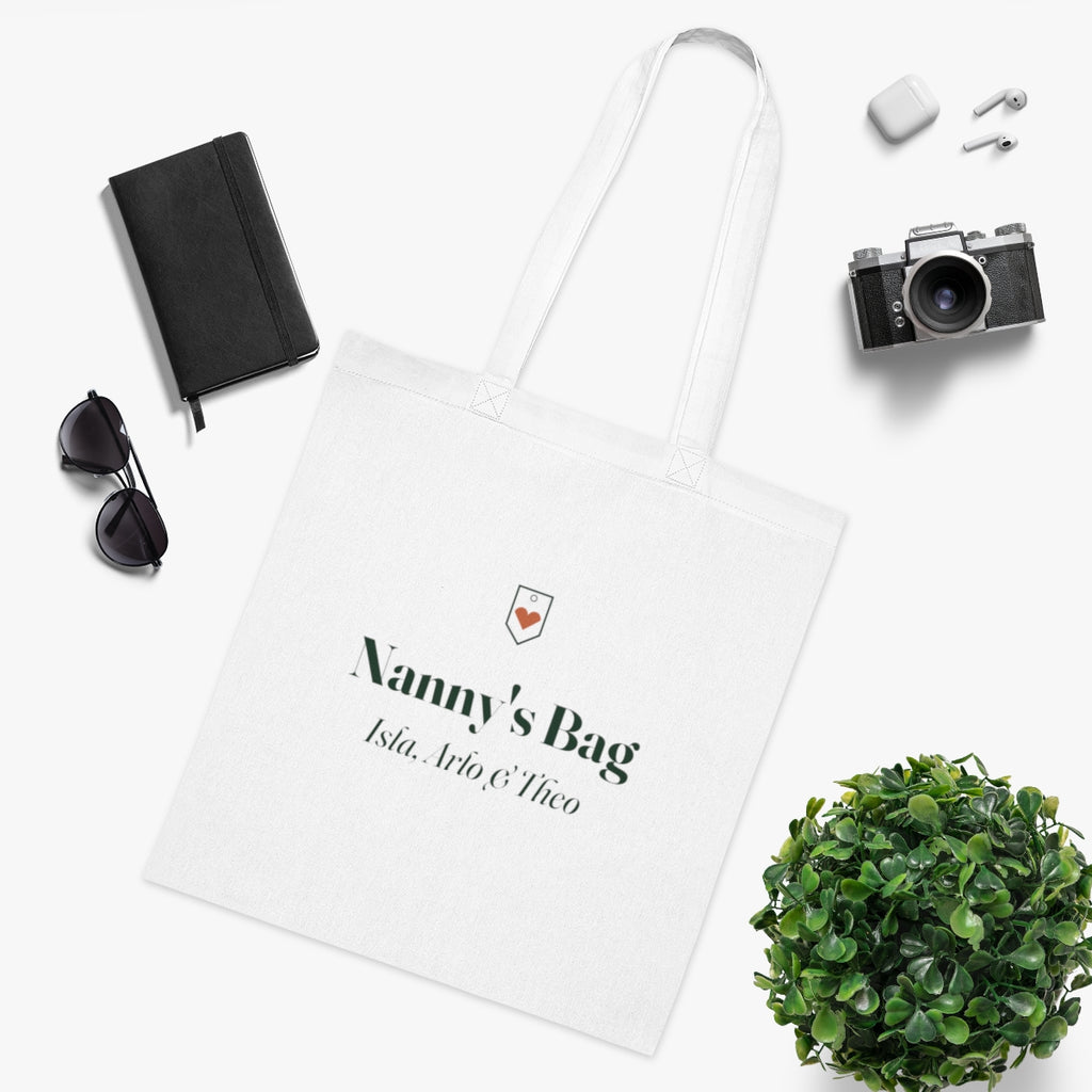 Personalised Nanny's Bag tote mock-up laid flat beside a notebook, sunglasses, earbuds, a camera and a leafy plant