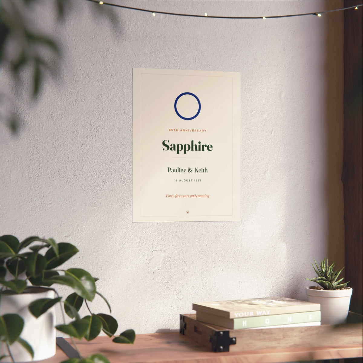 Sapphire anniversary print mock-up on a white wall above a desk with books, plants and fairy lights, larger size
