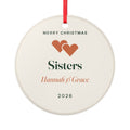 Sisters ceramic Christmas ornament mock-up, round with a red ribbon, two orange hearts, 'Hannah & Grace' and '2026'