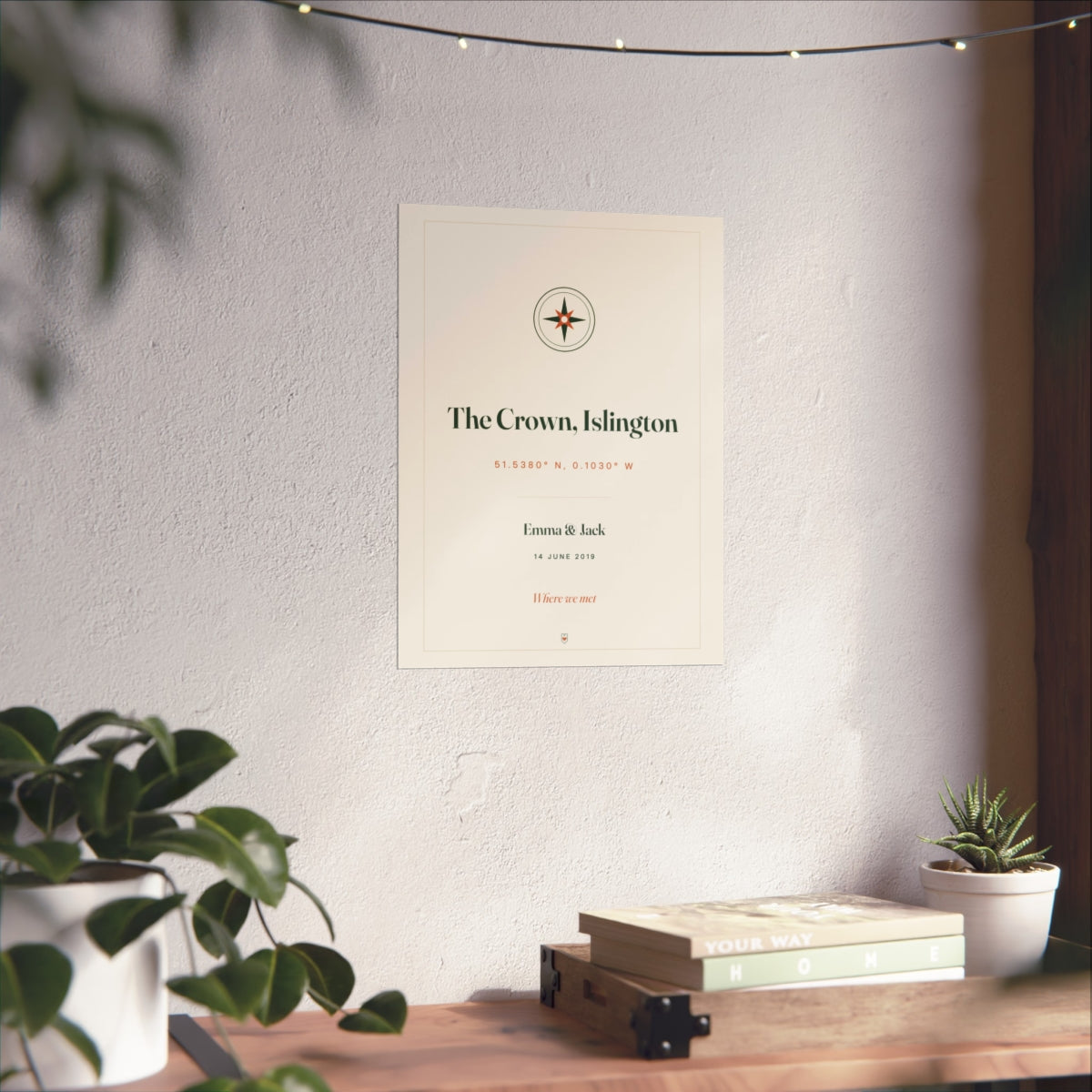 Personalised coordinates print mock-up on a white wall above a wooden desk with books and a small potted plant
